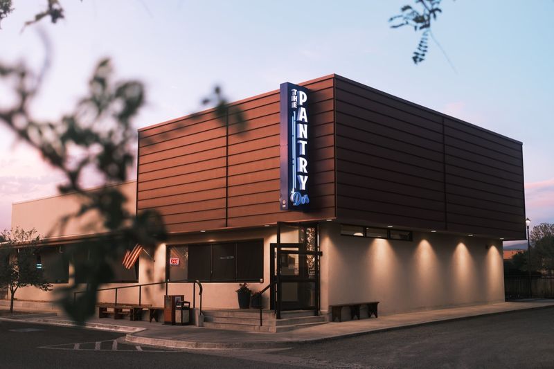 The Pantry Dos at dusk: the blue PANTRY Dos sign lit on the corner of the wood-clad building, lamps glowing along the wall.