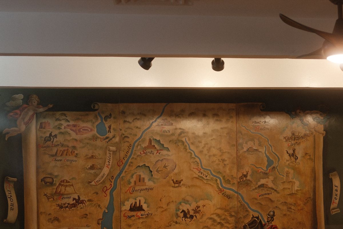 The top of the mural in the back dining room: a hand-painted map of New Mexico in aged yellows and greens, with the Rio Grande, Santa Fe, Taos and Albuquerque lettered in red, a cherub in each upper corner, and Arizona and Texas on scrolls at the edges, under track lights and a ceiling fan.
